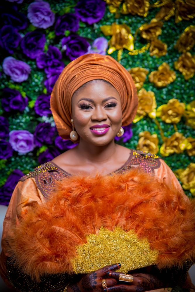 Bride in a gele holding an orange feather fan before a flower wall